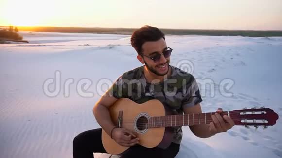 在夏季傍晚的露天日落时分在无尽的沙漠中坐着学弹吉他的穆斯林人的肖像视频的预览图