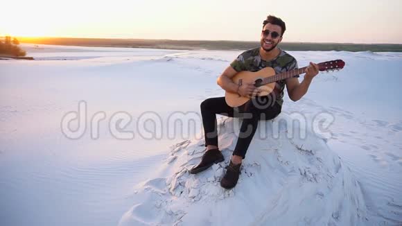 可爱的年轻男性阿拉伯人喜欢吉他的声音坐在沙漠中的山坡上日落时分在夏季的傍晚露天视频的预览图