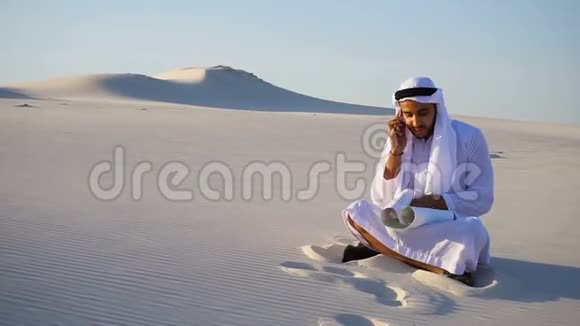 自信的男性阿拉伯阿联酋谢赫建筑工人与客户在手机上交谈坐在沙滩上用纸画大片视频的预览图