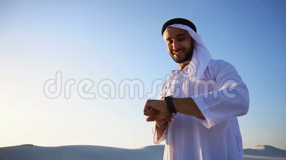 一幅英俊的阿拉伯商人的肖像他体验着新的智能手表站在炎热的阳光下广阔的沙漠中视频的预览图