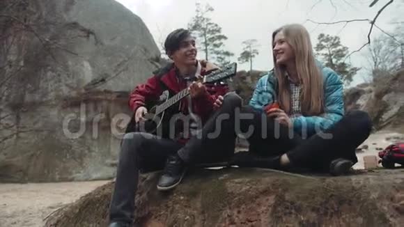 年轻人坐在岩石上唱歌年轻人弹吉他迷人的金发女孩喝热酒视频的预览图