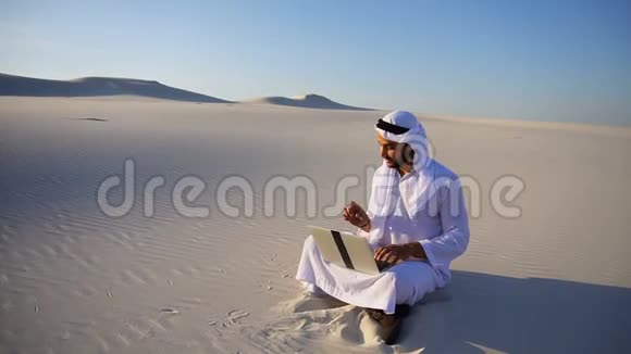 阿拉伯阿拉伯酋长国帅哥女商人在大沙漠里坐在沙滩上用笔记本电脑打电话给生意伙伴视频的预览图