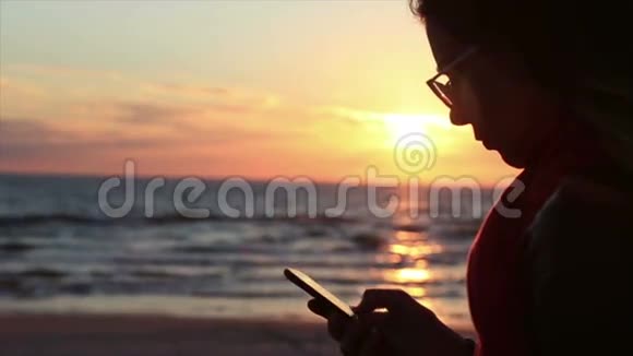 年轻的女孩子在海洋日落景观上使用智能手机视频的预览图
