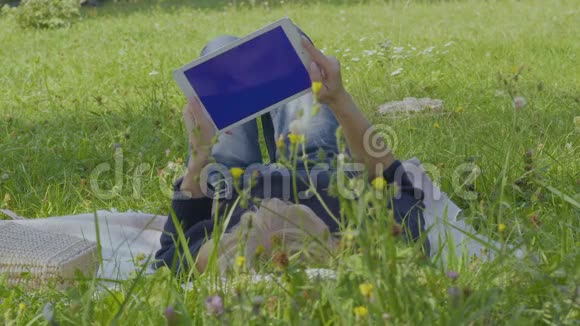 年轻女子用数码平板电脑在公园的毯子上放松视频的预览图