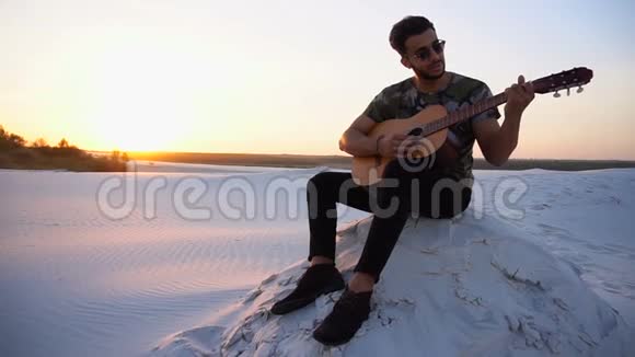 英俊的阿拉伯年轻人调整吉他坐在沙漠中的山上日落在温暖的夜晚露天视频的预览图