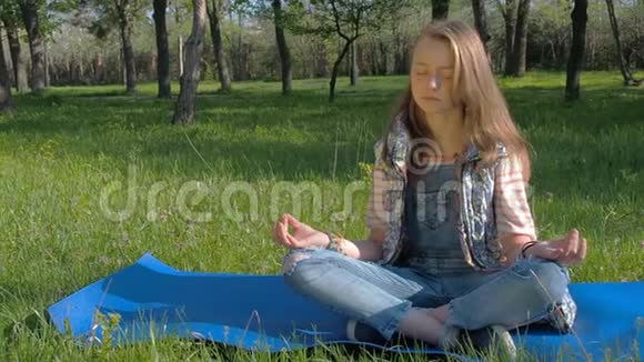 一个女孩坐在公园里做瑜伽姿势一个有雀斑的少女冥想一个穿牛仔裤的女孩在户外地毯上视频的预览图