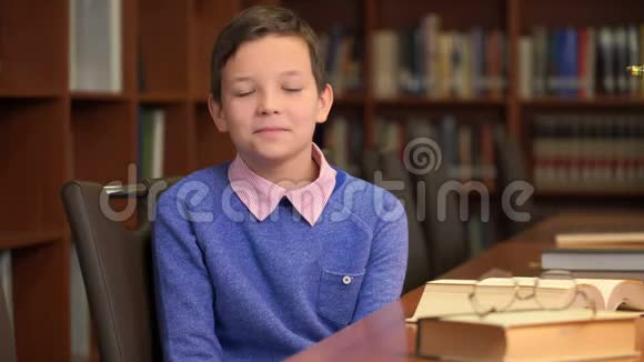 这位可爱的小学生坐在图书馆书架旁的画像视频的预览图