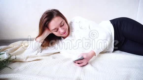 一幅可爱的女孩的肖像她在网上看新闻在舒适的房间里躺在格子布上一边高兴地看着视频的预览图
