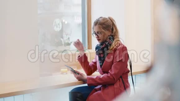 年轻的金发女孩使用她的设备在咖啡馆A触摸她的眼镜滑动触摸屏时尚的服装舒适视频的预览图