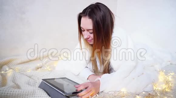 漂亮的女性看着平板电脑屏幕坐在互联网上躺在地板上下一个明亮的房间里穿着连衣裙视频的预览图