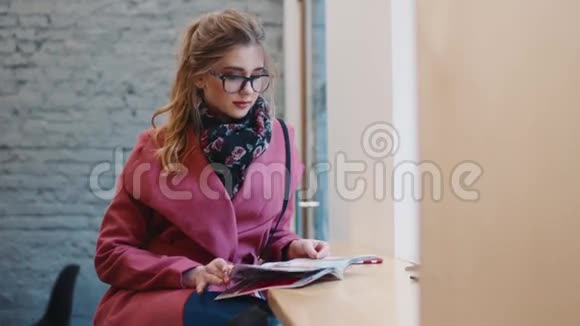 年轻迷人的女孩戴着时尚的眼镜独自坐在咖啡馆A读着时尚杂志时尚视频的预览图
