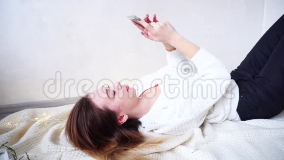 可爱的年轻女子在手机上打印信息躺在明亮的房间里的格子上挂着闪烁的花环视频的预览图