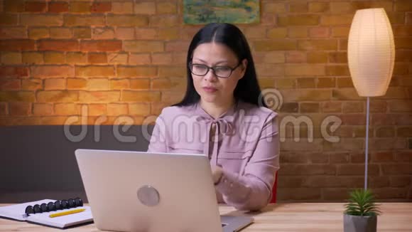 在办公室里一位成年亚洲女商人在笔记本电脑上打字看着摄像机微笑着视频的预览图