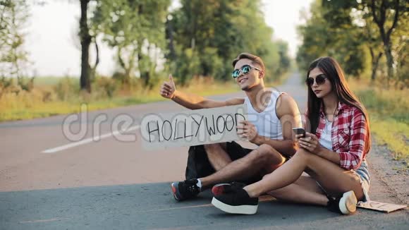 一对年轻夫妇坐在路上搭便车使用智能手机一男一女在高速公路上停车视频的预览图