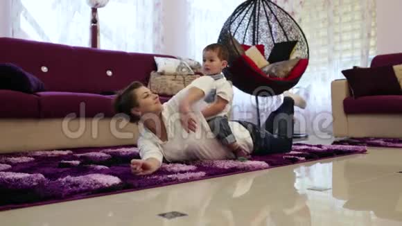 一位年轻的母亲和她的小儿子躺在客厅的地板上看电视玩得开心视频的预览图