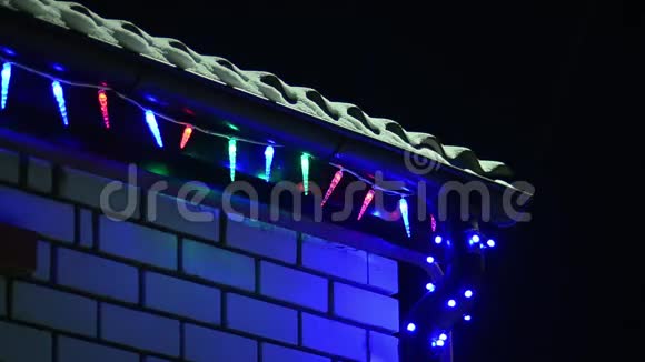 多种颜色的圣诞灯在晚上装饰房子雪躺在屋顶上闪闪发光的花环在冬天的屋顶上视频的预览图