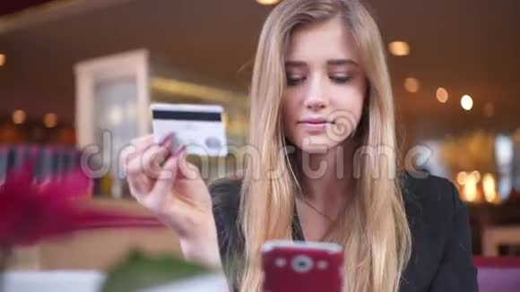 穿黑色夹克的可爱女孩在咖啡馆A她在智能手机上的银行卡数据方面处于领先地位视频的预览图
