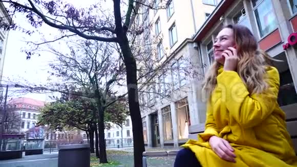 在凉爽的秋日坐在城市的长椅上坐在房子的背景下通过手机说话的美丽女孩的肖像视频的预览图