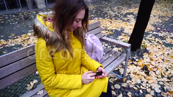 迷人的女性手持智能手机和浏览照片秋天下午坐在城市的长凳上视频的预览图