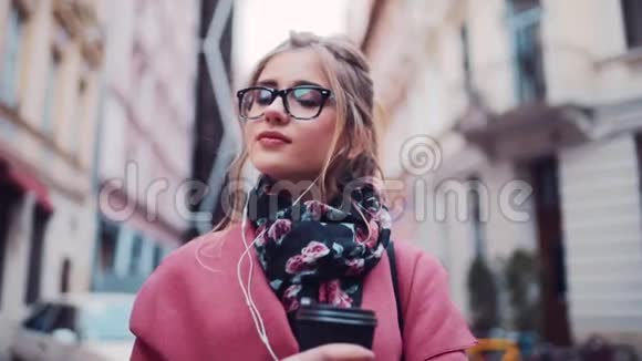 梦中的年轻女孩站在城市的街道上端着一杯咖啡用耳机听音乐看着很对视频的预览图