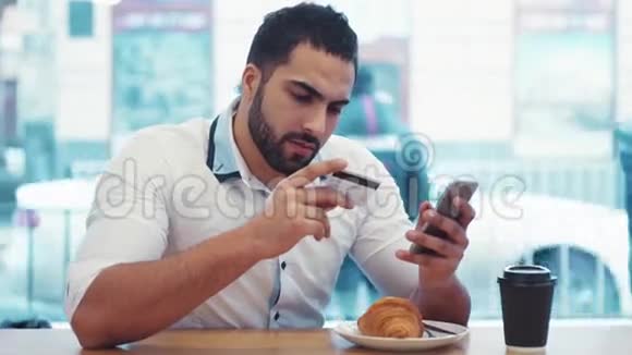 一位年轻的商人在A咖啡馆里坐着吃早餐时用智能手机使用网上银行购买视频的预览图
