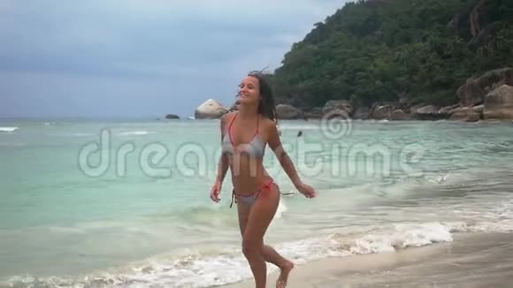在多云的天气里穿比基尼的年轻棕褐色漂亮女人在热带海滩海边奔跑慢动作1920x1080视频的预览图