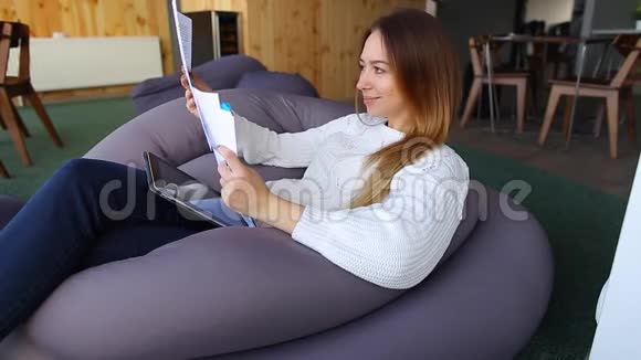 一位年轻可爱的女士的肖像她在春天的咖啡馆里学习文件快乐微笑坐在舒适的椅子上视频的预览图