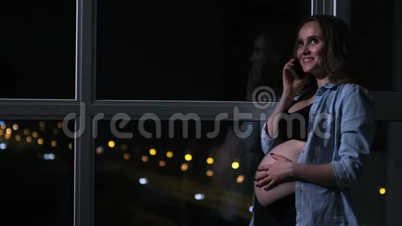 一个孕妇在夜里用手机说话手摸着站在窗边的肚子视频的预览图