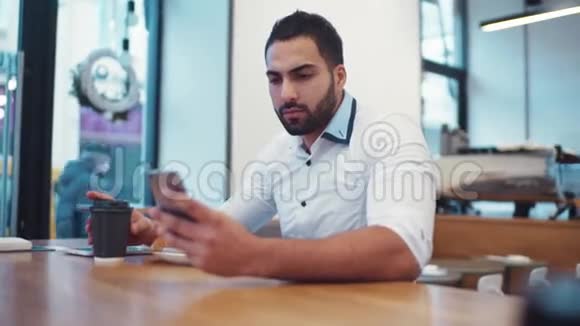 穿着白色正式衬衫的长胡子帅哥在喝咖啡时用手机上网现代视频的预览图