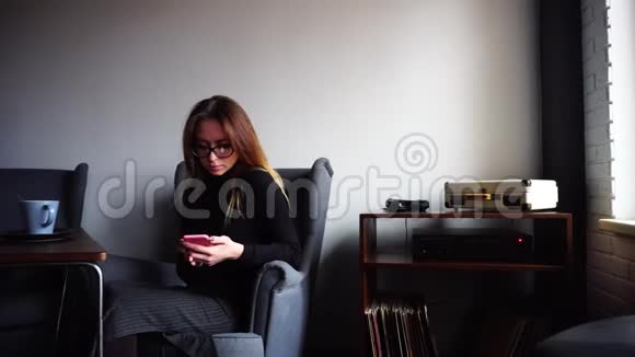 漂亮的女性在手机上打印信息看着相机微笑着坐在时尚办公室的灰色扶手椅上视频的预览图