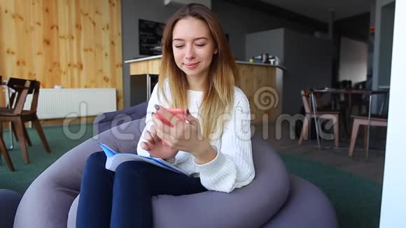 微笑的女人通过网络在手机上拨打Skype在秋天的一天坐在现代咖啡馆的软椅子上视频的预览图