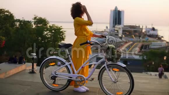 黎明时分一位身穿黄色长裙的漂亮黑发女子站在她的城市自行车旁手里拿着车把视频的预览图