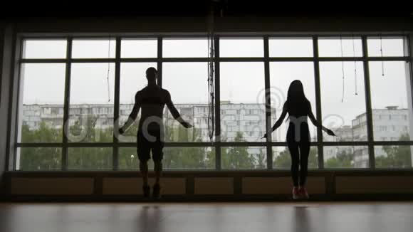 健身女性和肌肉男跳在靠近窗户的绳子上剪影视频的预览图