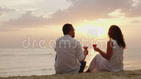 一对年轻夫妇在日落时正在海边喝酒他们坐在沙滩上戴着眼镜周年或蜜月视频的预览图