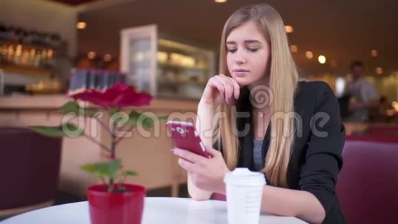 年轻的金发女孩穿着夹克坐在咖啡馆A商店她在用智能手机发短信视频的预览图
