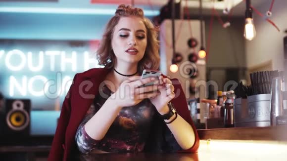 穿着红色外套的年轻女人独自坐在酒吧旁微笑着在手机里冲浪时尚的外观令人惊叹视频的预览图