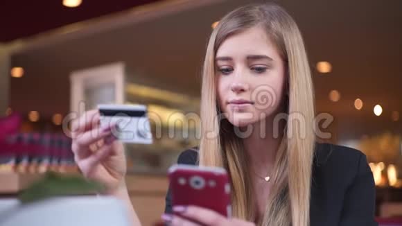 穿黑色夹克的金发美女在咖啡馆A她在智能手机上的银行卡数据方面处于领先地位视频的预览图