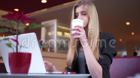 穿着黑色夹克的金发美女坐在咖啡馆里她正在喝咖啡用笔记本工作视频的预览图