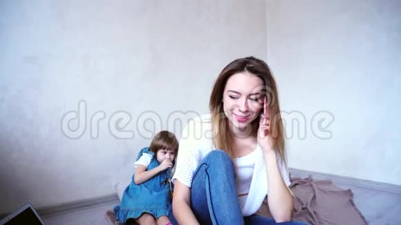 在明亮的房间里玩耍的小女儿的背景下可爱的女性和年轻的母亲在电话中交谈视频的预览图