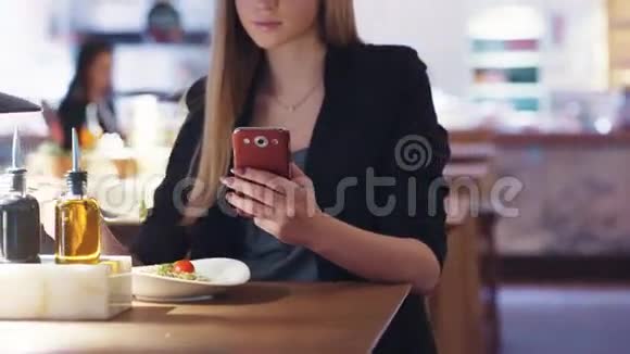 穿着黑色夹克的可爱的金发美女坐在咖啡馆A里她正在和一个朋友发短信吃午饭视频的预览图