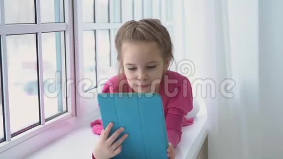 一个穿着粉红色裙子的小女孩正坐在窗边端着一块平板电脑视频的预览图
