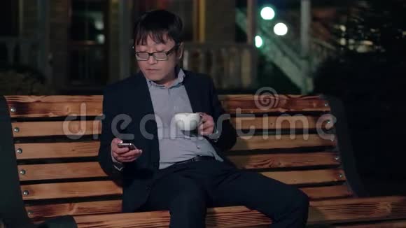 夜晚公园的亚洲商人使用智能手机喝一杯咖啡微笑视频的预览图