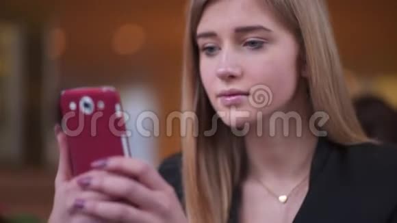 穿着黑色夹克的快乐可爱的女孩坐在咖啡馆A商店里她在用智能手机发短信视频的预览图