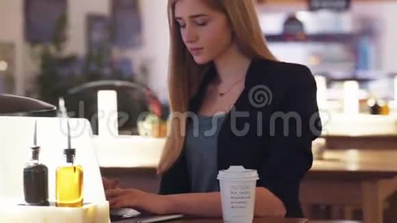 穿着黑色夹克的漂亮年轻女人坐在咖啡馆A商店里她用笔记本专注于工作视频的预览图