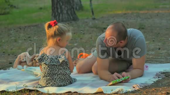 一家人在森林里的毯子上放松父亲教小女儿读书视频的预览图