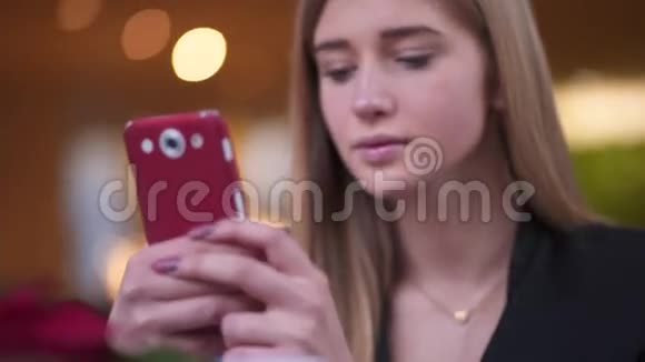穿着黑色夹克的微笑的年轻女孩坐在咖啡馆A商店里她在用智能手机发短信视频的预览图