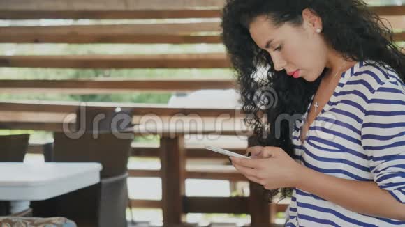 夏季露台上使用智能手机的美丽卷发黑发女郎视频的预览图
