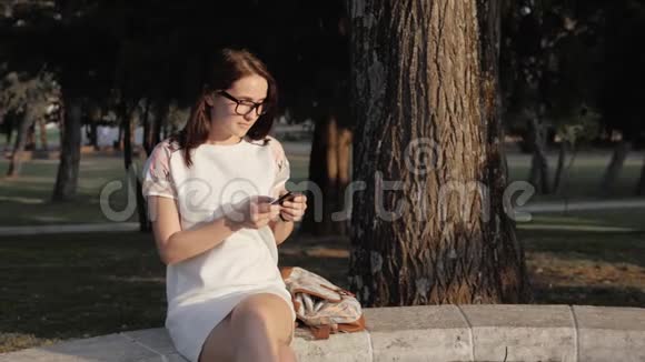 年轻微笑的女人在日落时坐在公园的长凳上用智能手机美丽的欧洲女孩在电话里发短信视频的预览图