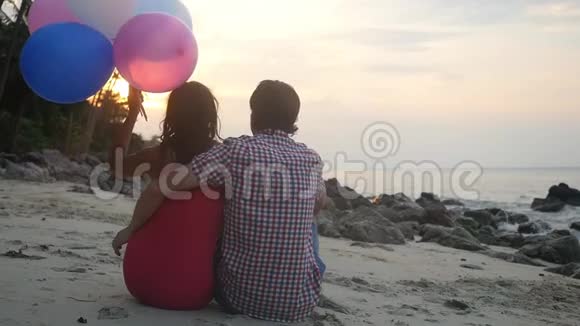 年轻夫妇坐在海滩上抱着气球看着日落慢动作1920x1080视频的预览图
