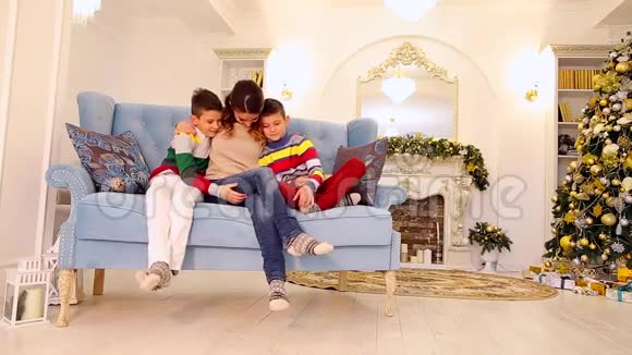 勤劳的母亲和两个双胞胎男孩和孩子并排坐在蓝色沙发上以家庭的方式拥抱在节日里视频的预览图
